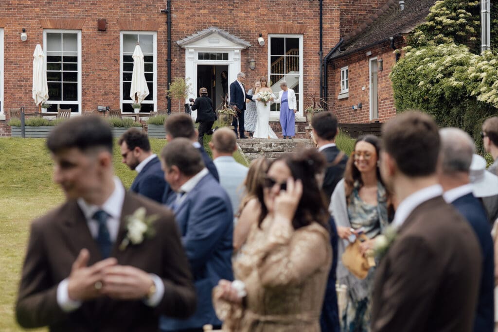 Bride exiting Stockton House and walking towards outdoor ceremony while guests gather on lawn in Shropshire