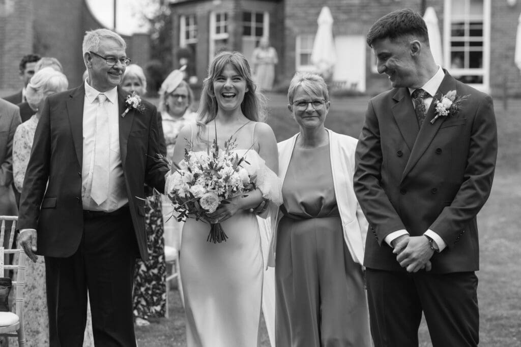 Bride holding bouquet and walking down aisle with her parents during outdoor wedding ceremony at Stockton House, Shropshire