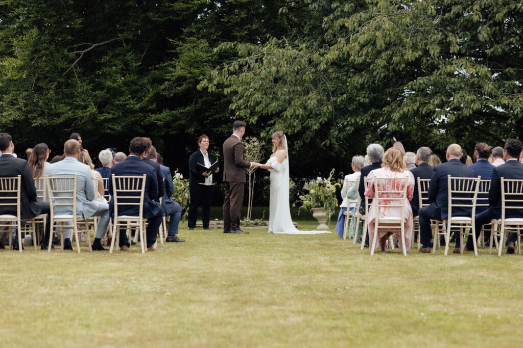 Bride and groom standing together during outdoor wedding ceremony on lawn at Stockton House, Shropshire with guests seated