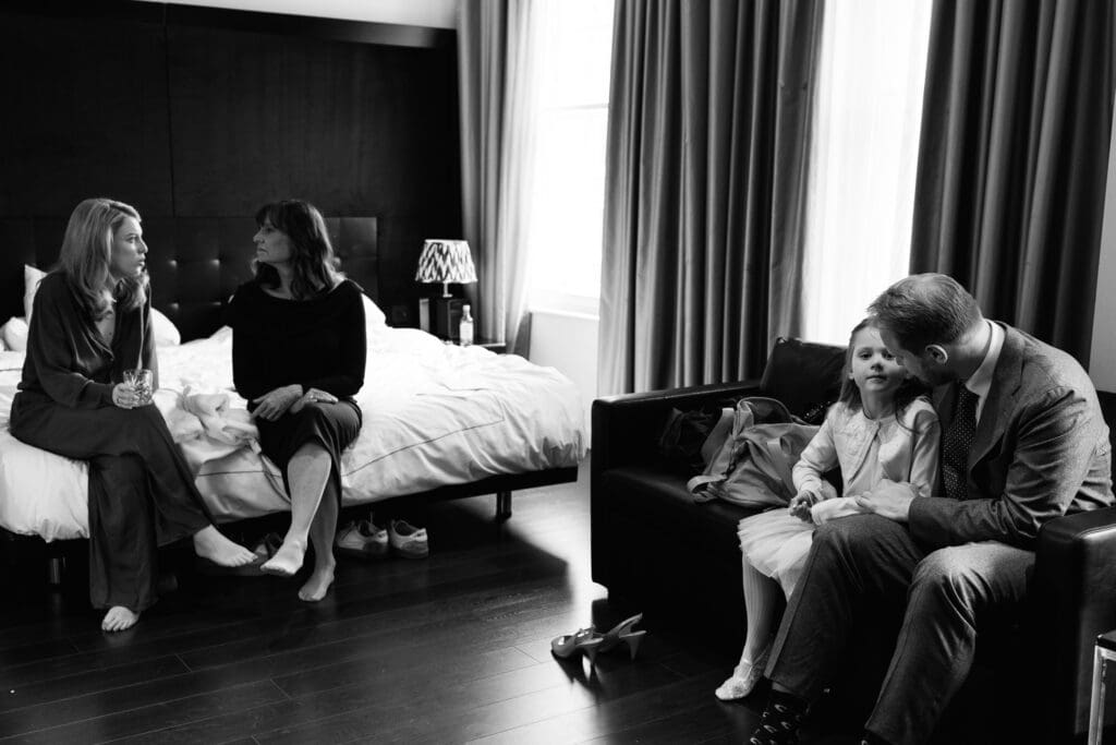 mother of the bride and bride are sat on the bed in the background deep in conversation while the groom and his daughter are speaking together on the sofa right of frame