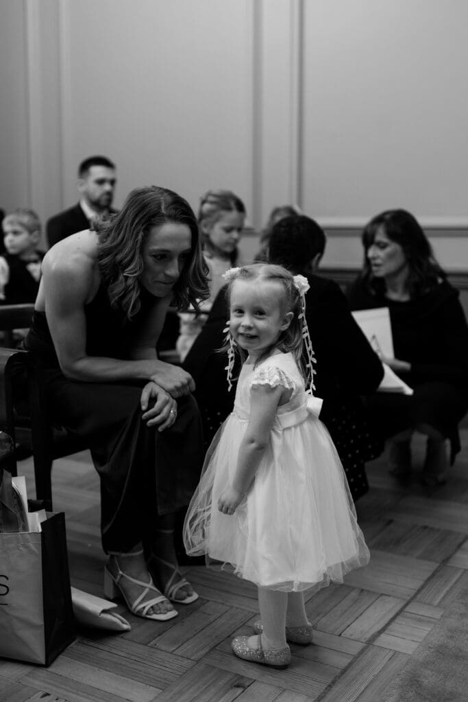 a young girl smiles at the camera as one of the bridesmaids tries to talk to her and get her attention just before the wedding ceremony at Marylebone Town Hall in London