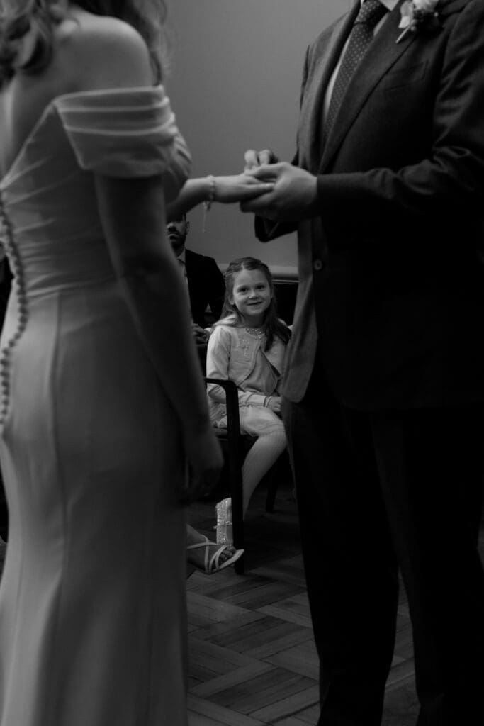 the grooms daughter looks at her father and also her step mother as they exchange rings. The daughter is framed by the couple's hands in the Knightsbridge Room in Marylebone Town Hall in London