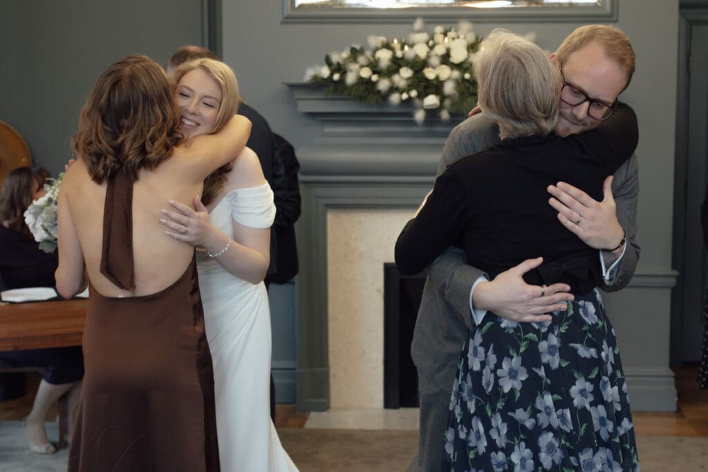 the bride and groom hug relatives balancing the frame in a double hug in the Knightsbridge room at Marylebone Town Hall in London after their wedding ceremony