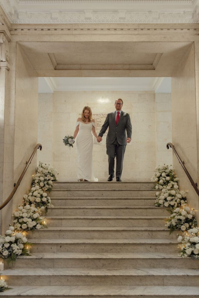 the bride and groom make their way down the flower lined iconic interior staircases after their wedding ceremony at Marylebone Town Hall in London