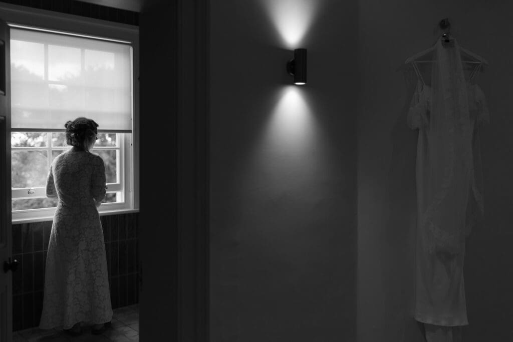 Bride looking out of window during morning preparations with wedding dress hanging nearby at Stockton House, Shropshire