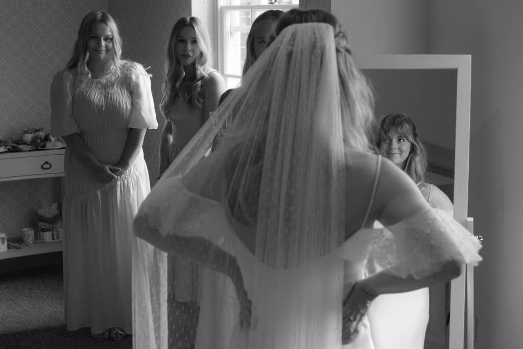 Bride in wedding dress and veil facing mirror while bridesmaids look on during morning at Stockton House, Shropshire