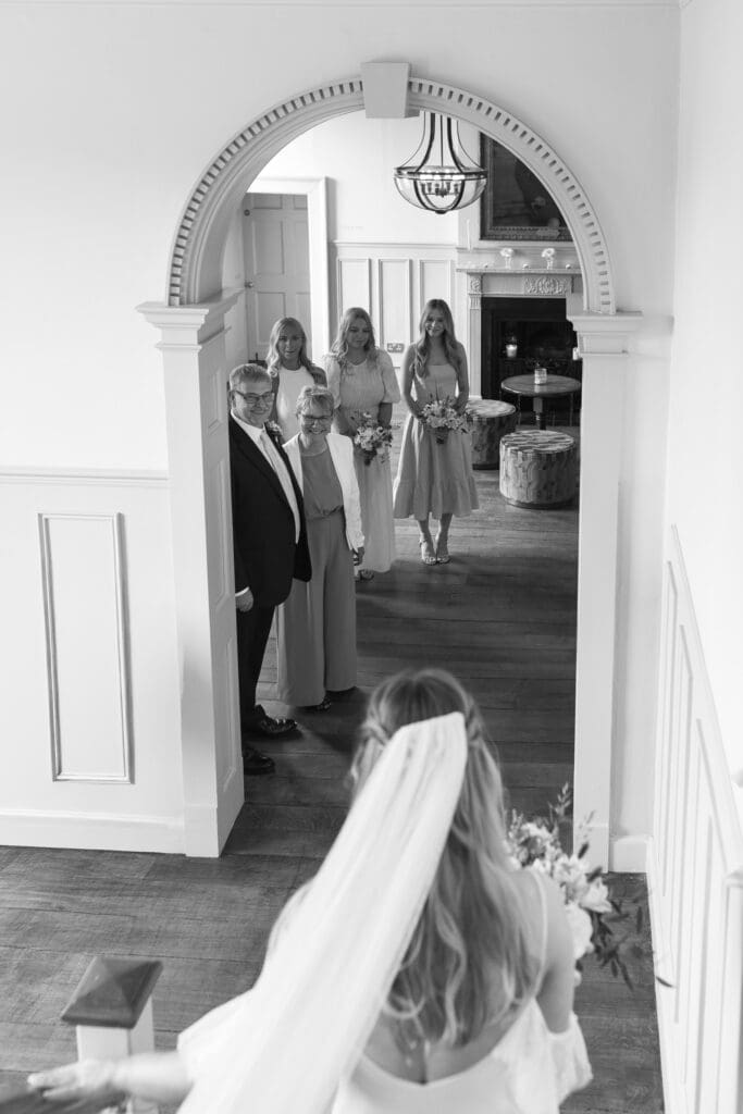 Bride in veil walking down staircase towards family and bridesmaids waiting under archway at Stockton House, Shropshire