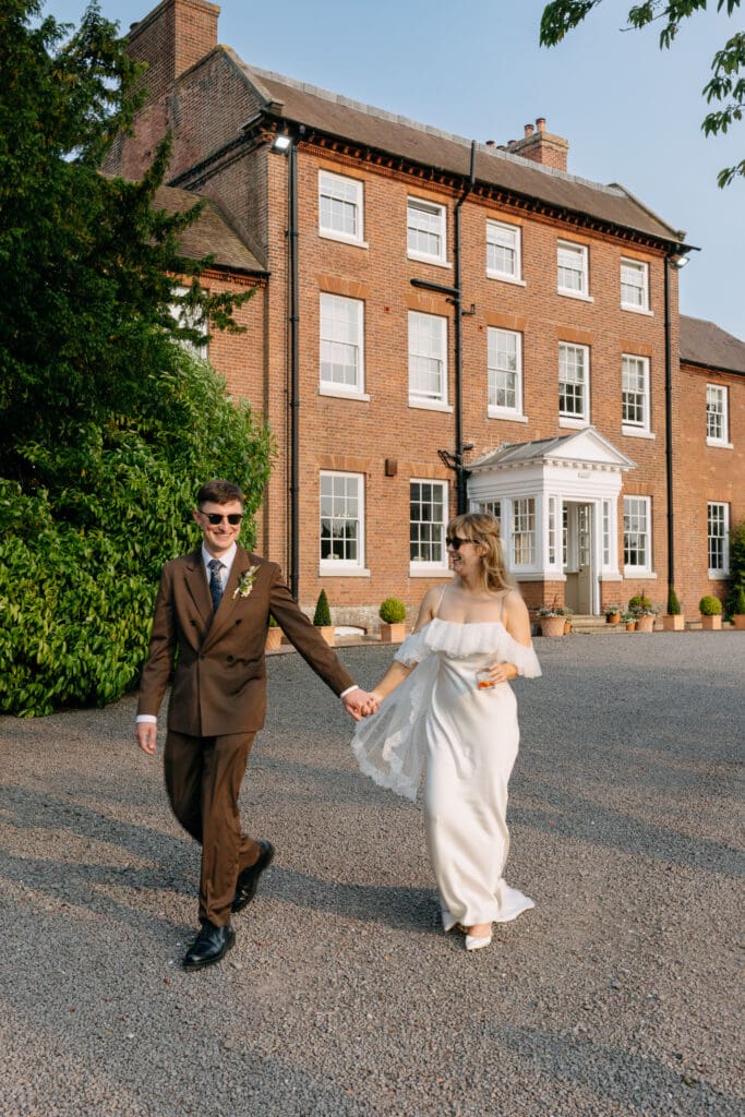 Bride and groom holding hands and walking outside Stockton House, Shropshire in warm evening light