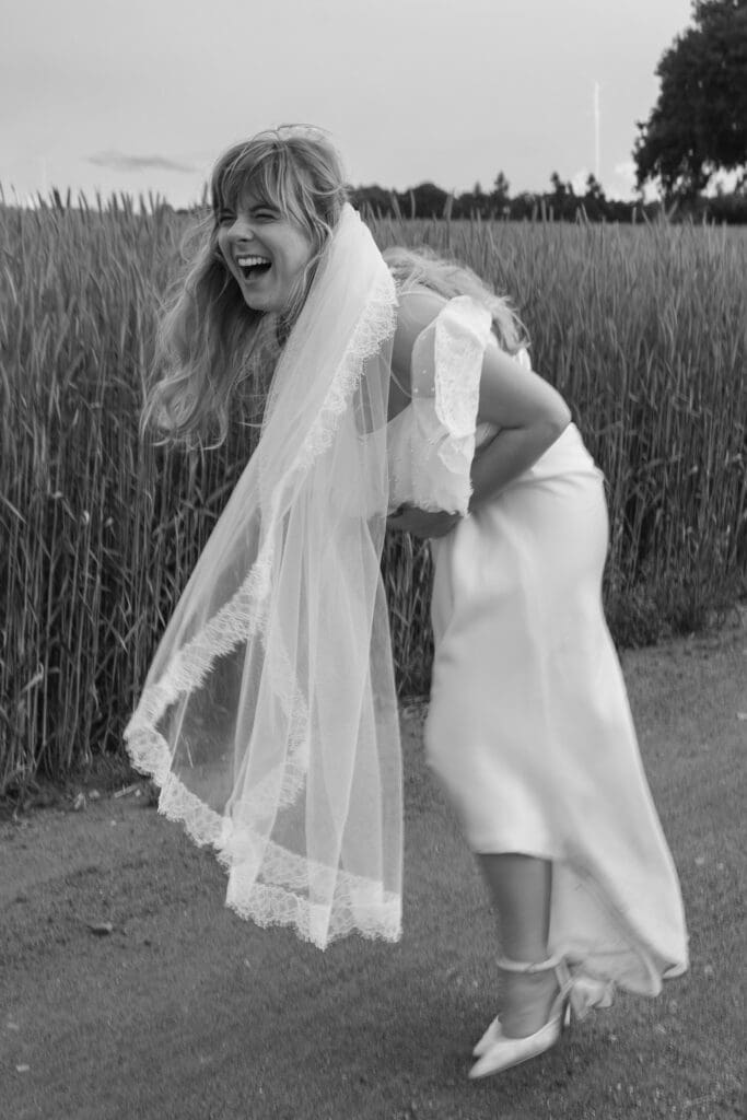 Bride in wedding dress and veil laughing while bending forward during couple portraits in field at Stockton House, Shropshire
