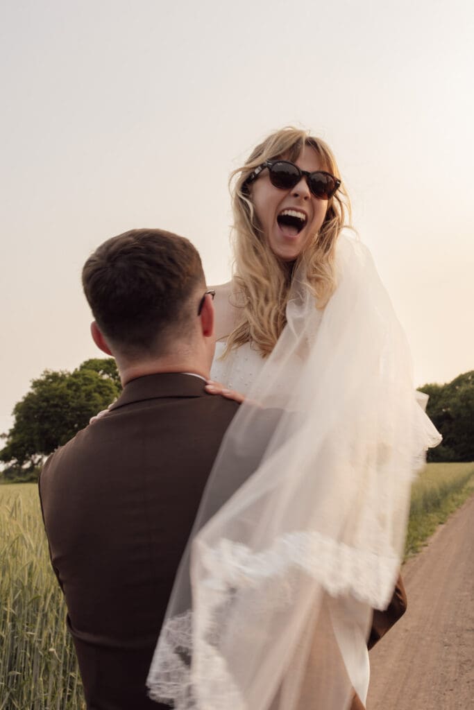 Bride wearing sunglasses being lifted by groom during golden hour couple portraits in countryside near Stockton House, Shropshire