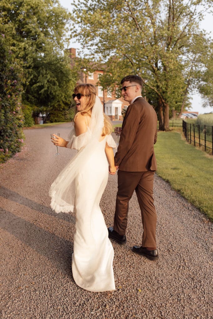 Bride and groom holding hands and walking outside Stockton House, Shropshire in golden hour light, both wearing sunglasses
