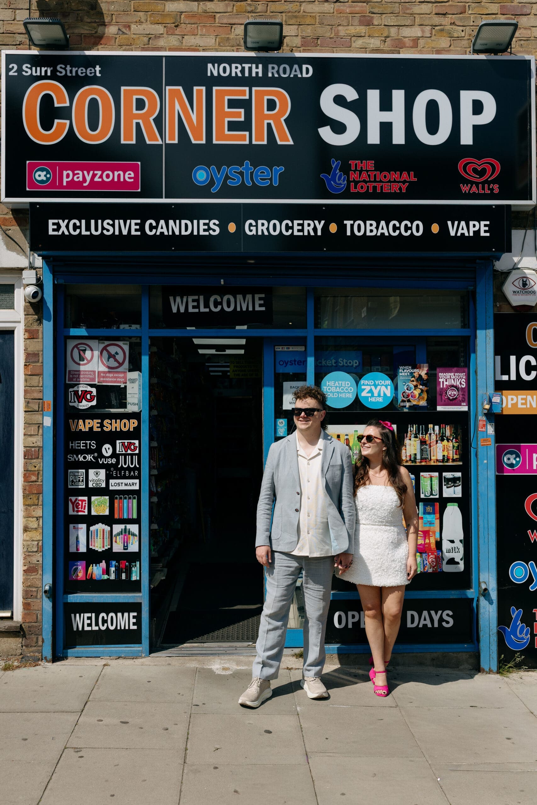 Becca and Dom standing outside a London corner shop during wedding portraits.